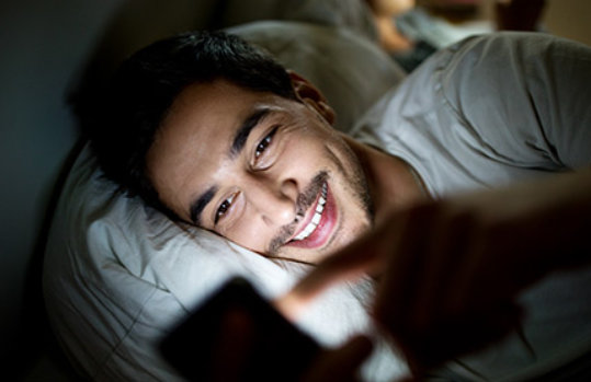 Smiling man uses mobile device while in bed at night