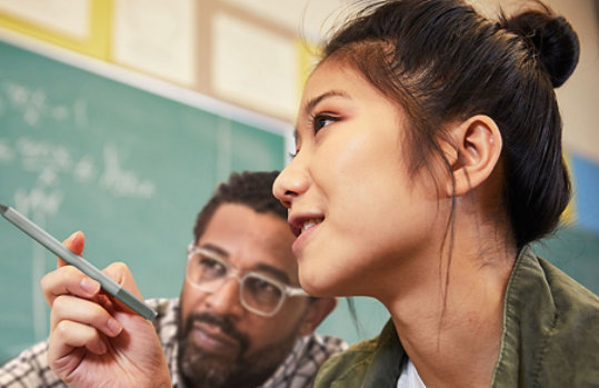 Female student points to her work, explaining to a peer. Set in a classroom environment.