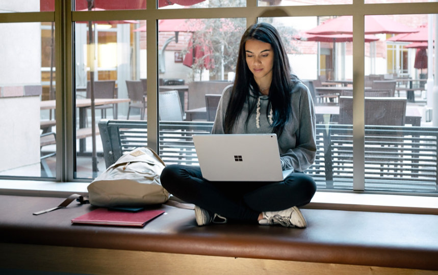 Uma mulher jovem sentada com as pernas cruzadas em um parapeito usando um Surface Book no colo.