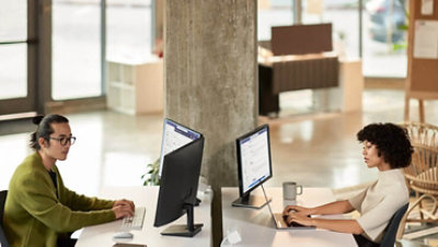 A man and a woman collaborating during a Microsoft Teams meeting while working in an open office setting on dual monitors.