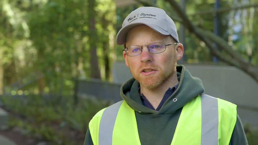 Kyle is speaking, standing outside. There are trees and landscaping behind him. He is wearing a ball cap and a high visibility vest.