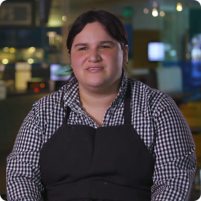 A Supported Employee, Nicole, is seated talking. She is wearing a black and white gingham shirt and a black apron.