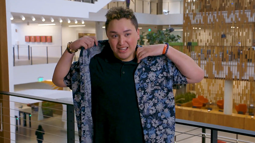 Austin is smiling, putting on his blue flowered Hawai'ian shirt. In the background is a corporate office setting.