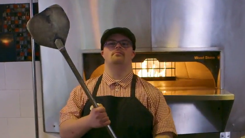 Phillip is standing in a cafe holding a pizza peel. There is fire visible in a woodfire stove behind him. He is wearing a ball cap, and red shirt and an apron.