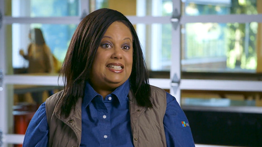 Leila is seated, smiling, in an office setting with large windows and plants in the background. She wears a blue button-up shirt and khaki waistcoat.