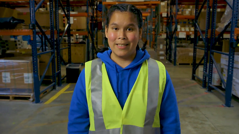 Tanya is smiling, standing in a warehouse. She is wearing a blue hoodie and a high visibility vest. Racks with boxes and other materials are in the background of the warehouse.