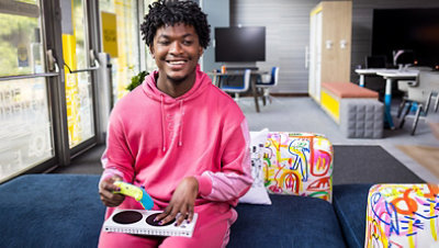 A black woman with short hair, slight beard, and a wonderful smile sits on a brightly colored couch, in a pink tracksuit, holding a brightly colored Xbox controller and an Xbox Adaptive Controller on her lap.