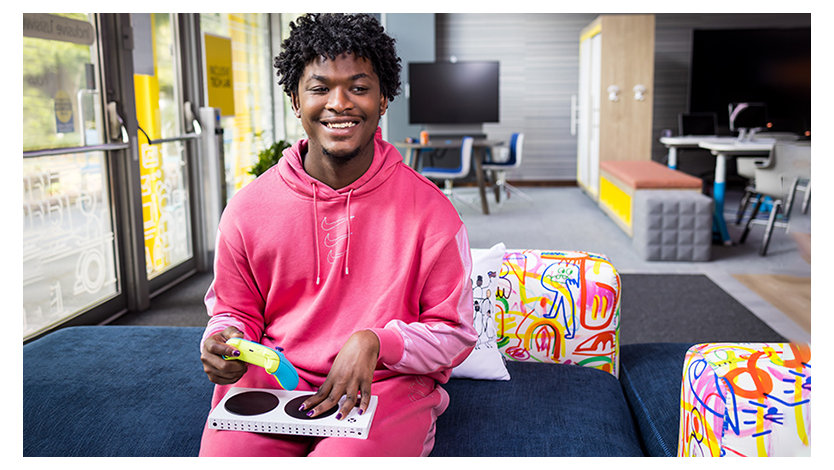 A black woman with short hair, slight beard, and a wonderful smile sits on a brightly colored couch, in a pink tracksuit, holding a brightly colored Xbox controller and an Xbox Adaptive Controller on her lap.