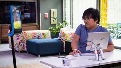 An Asian man with thick glasses sits at a desk in front of a Surface Pro mounted on a stand at eye level he's talking to an assistive robot used for telecommuting.