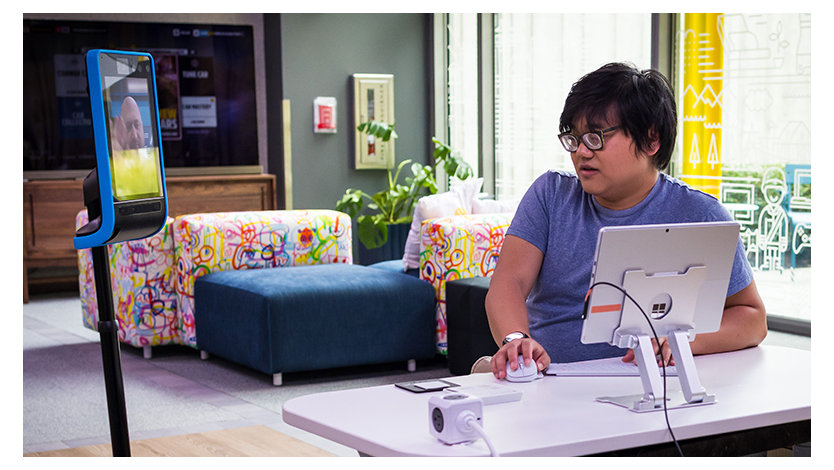 An Asian man with thick glasses sits at a desk in front of a Surface Pro mounted on a stand at eye level he's talking to an assistive robot used for telecommuting.