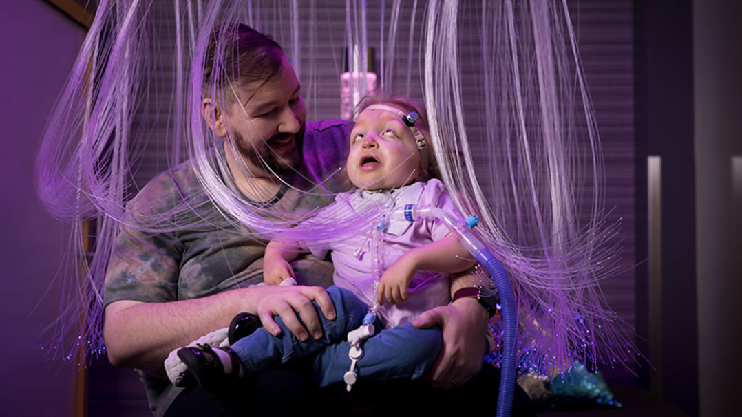 In a colorful cascade of fiber optic lights, a man holds his daughter, a small for her age 4-year-old disabled girl with craniofacial differences. She uses a hearing-aid and breathes using a ventilator. 