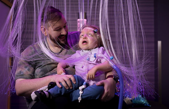 In a colorful cascade of fiber optic lights, a man holds his daughter, a small for her age 4-year-old disabled girl with craniofacial differences. She uses a hearing-aid and breathes using a ventilator. 