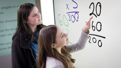 Two young women work at a Surface Hub on math problems the woman in the foreground is pointing at a multiplication problem with her limb different hand.