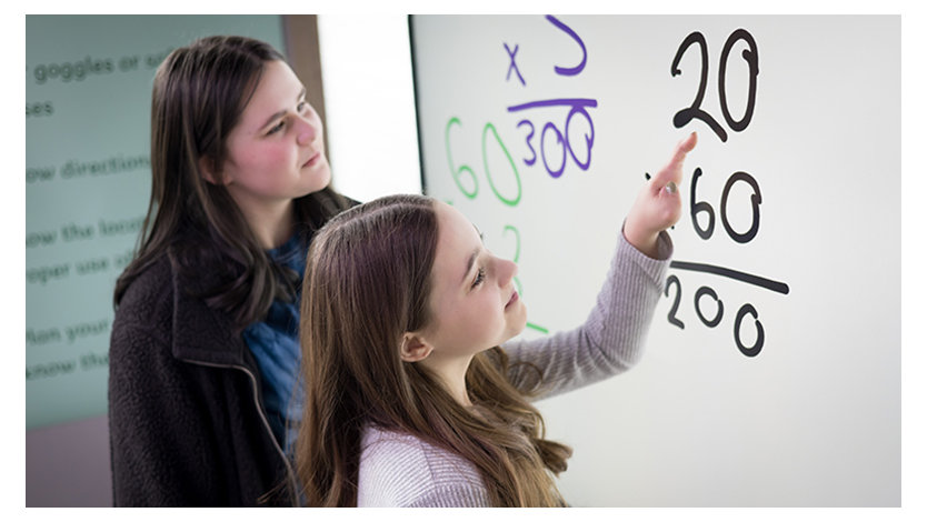Two young women work at a Surface Hub on math problems the woman in the foreground is pointing at a multiplication problem with her limb different hand.