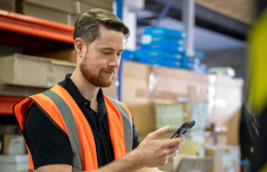 Man wearing an orange vest in a manufacturing or warehouse who appears to be doing inventory or looking at something in a box and messaging about it on his phone in Microsoft Teams.