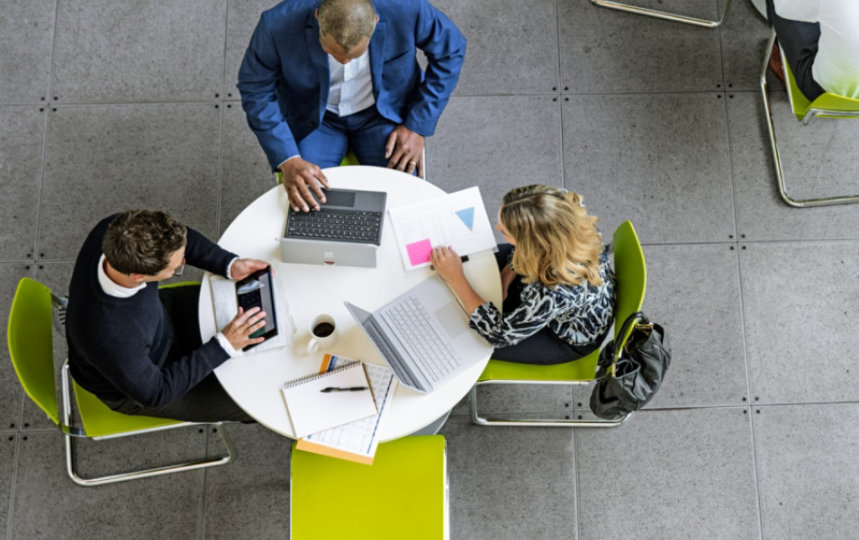 Top-down view of two men and one woman gathered around round white table, using laptops and a tablet.