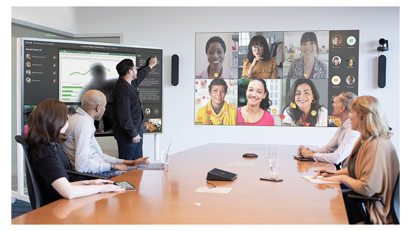 Five people are hosting a coordinated meeting in a large conference room and are joined by several virtual attendees on a projection screen in Front of Room view.