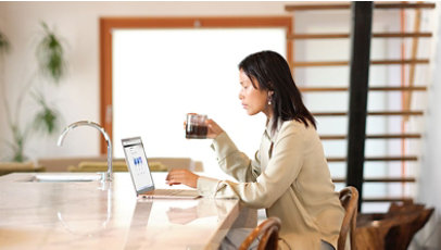 Woman sitting in kitchen, drinking coffee, while looking at laptop screen  