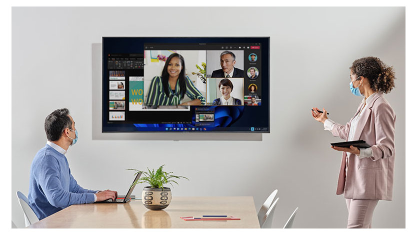 Two people in a room wearing masks looking at a screen with several Teams meeting attendees