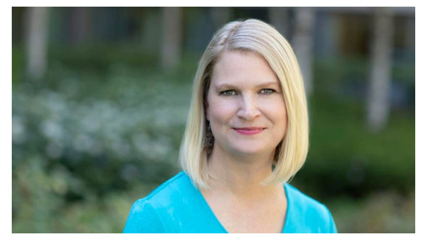 A photograph of Lindsay-Rae McIntyre wearing a blue sweater standing in front of greenery.