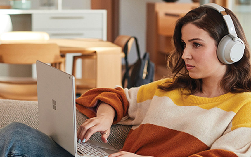 A woman uses a Surface laptop and Surface Headphones while lounging on a couch at home.
