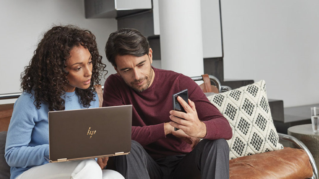 Man and woman sitting on a couch, looking at a mobile device and Windows 10 laptop