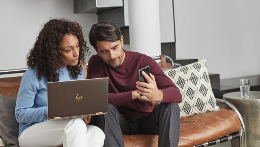 Man and woman sitting on a couch, looking at a mobile device and Windows 10 laptop
