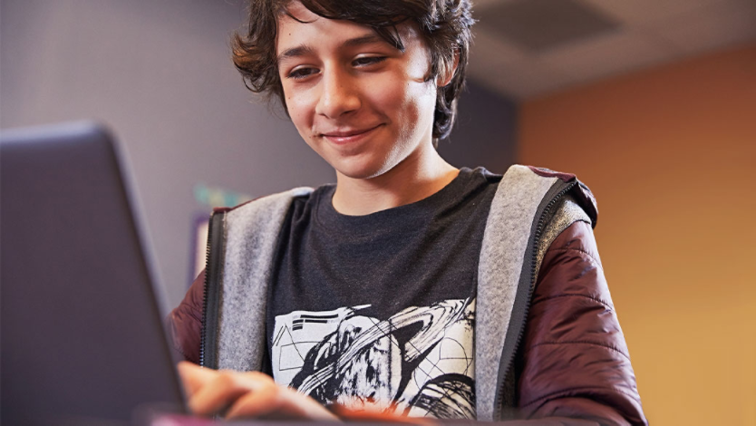 Male middle school student grins while using laptop, seated at desk in bedroom