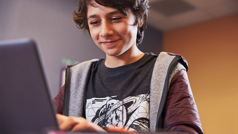 Male middle school student grins while using laptop, seated at desk in bedroom.