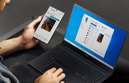 Man holding a Samsung mobile phone next to a laptop computer showing the Microsoft Your Phone app