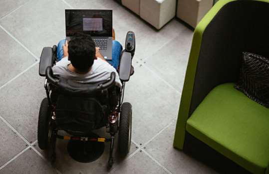 Person using a wheelchair, works on a laptop while in the lobby of an office building.