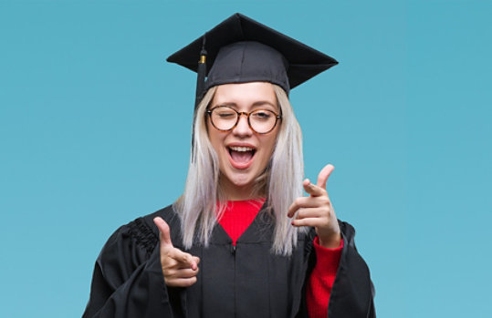 Photo credit: Aaron Amat/iStock/Getty Images. Young woman wearing graduation gown and pointing fingers and winking at camera