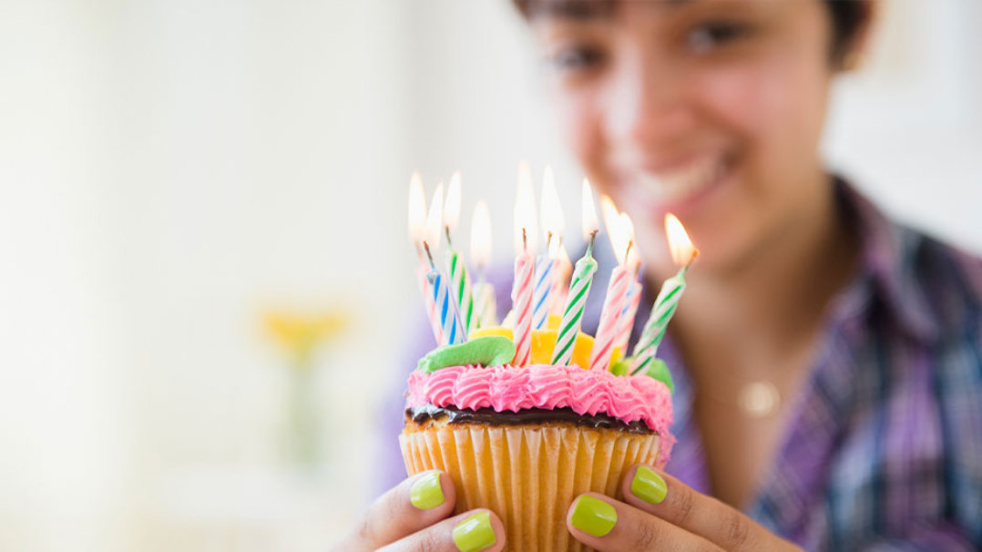 Photo credit: JGI/Jamie Grill/Getty Images. A smiling woman holding a cupcake with colorful birthday candles