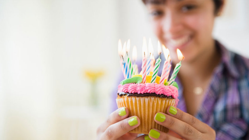 Photo credit: JGI/Jamie Grill/Getty Images. A smiling woman holding a cupcake with colorful birthday candles.