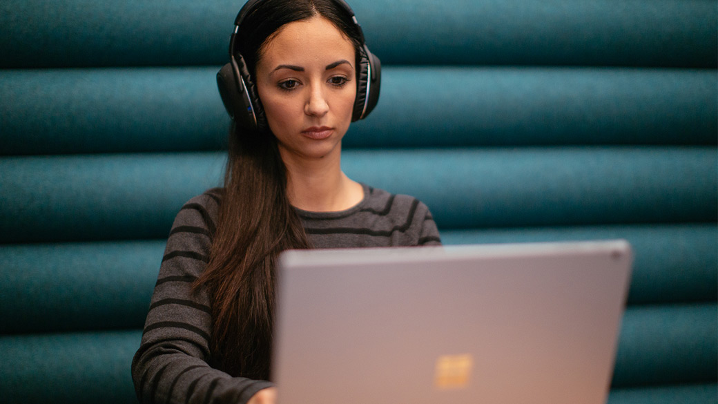 Woman wearing headphones is seated at a small table and types on a Surface computer.