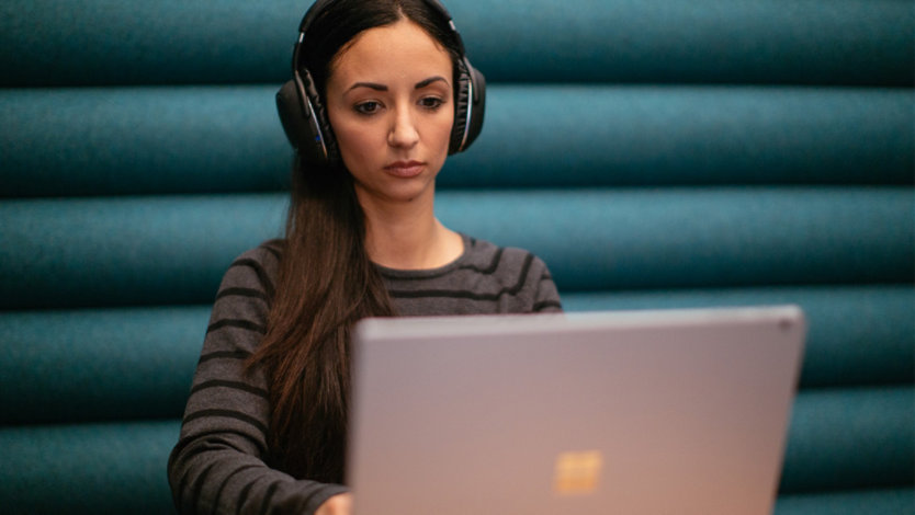 woman wearing headphones is seated at a small table and types on a Surface computer