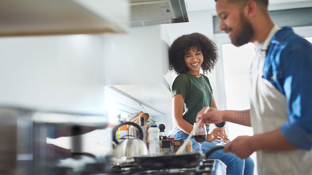 Smiling woman sitting on kitchen counter with a glass of wine watches man cook food on a stovetop