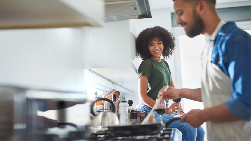 Photo credit: LaylaBird/E+/Getty Images. Smiling woman sitting on kitchen counter with a glass of wine watches man cook food on a stovetop