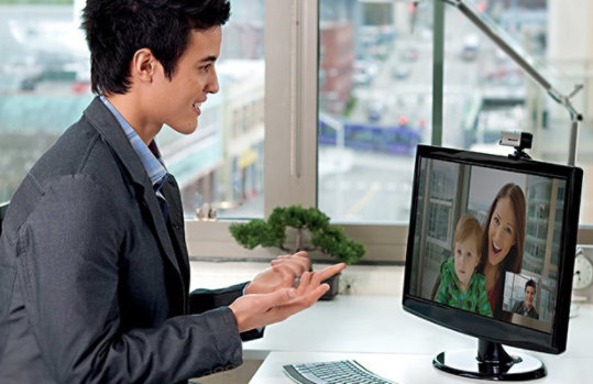Homem sentado em uma mesa olhando para um monitor com uma webcam LifeCam Studio encaixada nele
