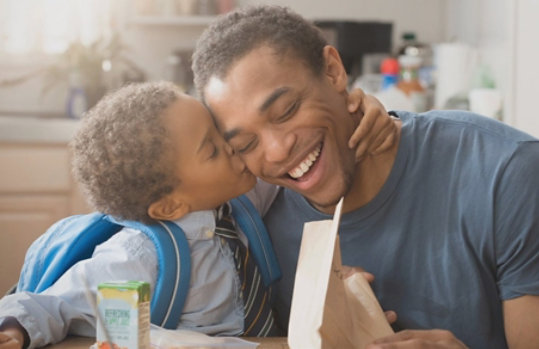 Young son embraces father, kissing his cheek, while sitting in a kitchen