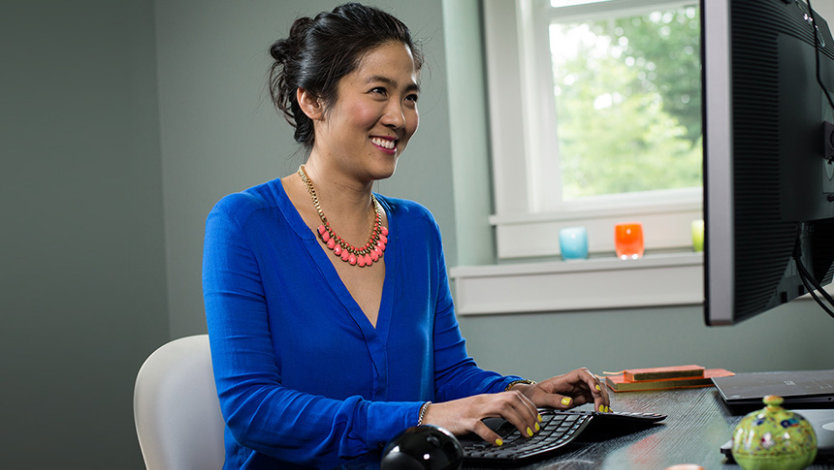 O femeie stă la birou, zâmbind către monitor în timp ce tastează la tastatură