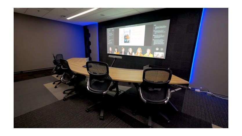 Meeting room with a table curving around and facing a wall display where virtual attendees and meeting content are shown together.