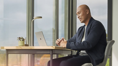 A person sitting at a desk with their hands folded participating in a video call on a laptop. 