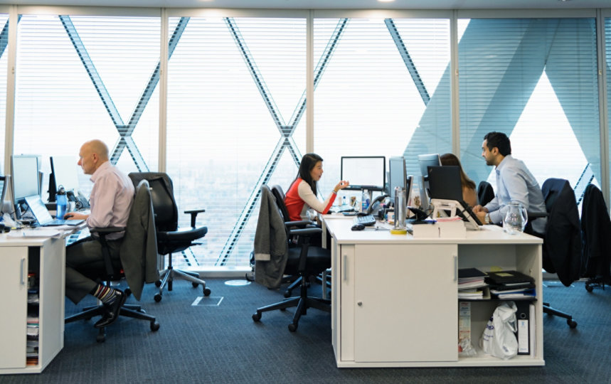 A female and male office worker in conversation, facing each other at shared workstation in open office setting. 