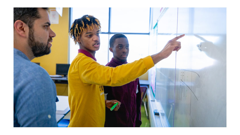 Two young boys with their teacher pointing at something on a whiteboard.