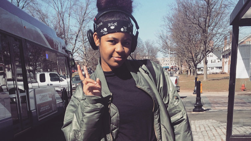 A young woman standing outside near at a bus stop wearing headphones and giving a peace sign with her right hand.