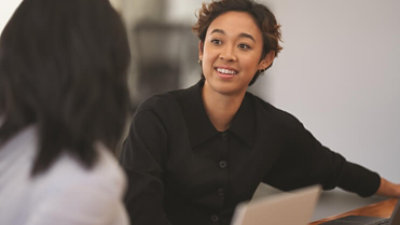 A woman smiles as she talks to another Microsoft employee while sitting a desk in an open working space.