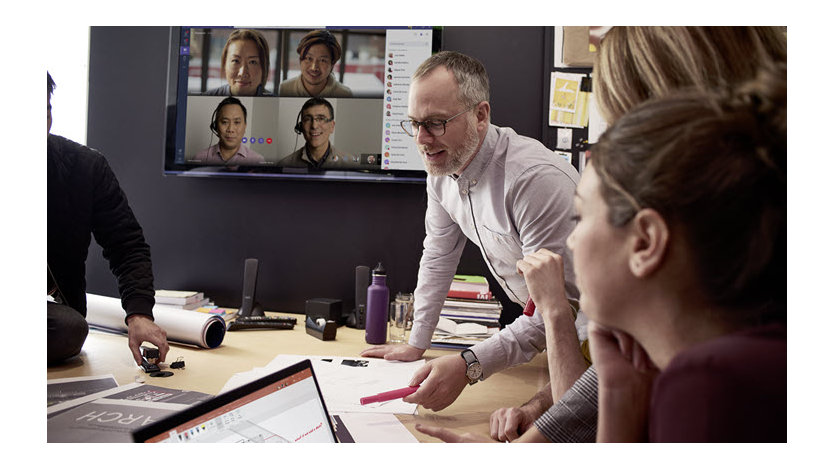 One person speaks at a conference room table as four people look on via a screen on Microsoft Teams and several others watch from in the room.