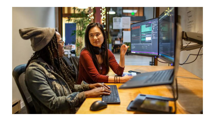 Two women talk to each other in front of computer screens with code displayed.