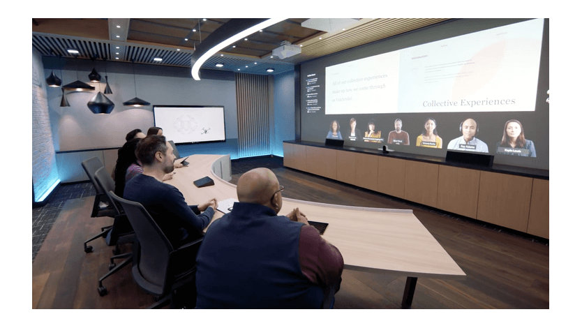 Microsoft employees are shown at a curved table facing a screen as people in the room and joining remotely meet.